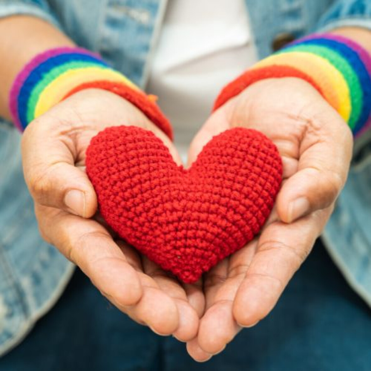 A pair of hands, with rainbow wristbands is showing a red crocheted heart to the screen